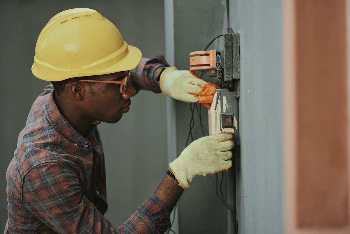 An electrician in a hard hat and gloves working on a meter box